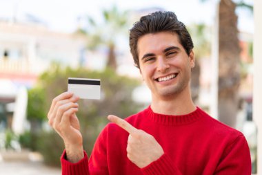 Genç yakışıklı bir adam dışarıda kredi kartı tutuyor ve onu doğrultuyor.