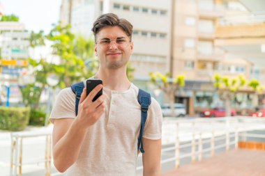 Genç yakışıklı adam dışarıda cep telefonu kullanıyor, üzgün bir ifadeyle.