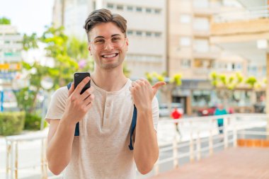Dışarıda cep telefonu kullanan yakışıklı genç bir adam bir ürün sunmak için yan tarafı işaret ediyor.