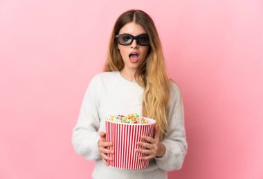 Pembe arka planda izole edilmiş genç sarışın kadın 3D gözlüklü ve elinde koca bir kova patlamış mısırla şaşırdı.