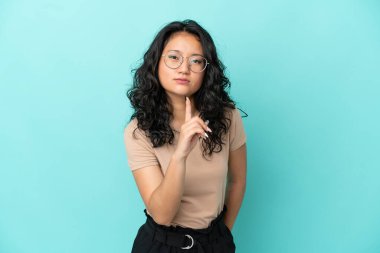 Mavi arka planda izole edilmiş genç Asyalı kadın hayal kırıklığına uğramış ve ön tarafı işaret ediyor.