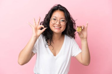 Pembe arka planda Bitcoin tutan genç Asyalı kadın parmaklarıyla 