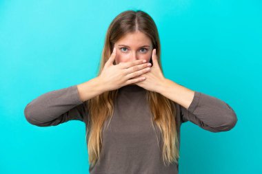 Mavi arka planda izole edilmiş genç sarışın kadın elleriyle ağzını kapatıyor.