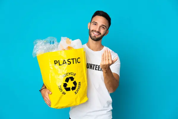 Young caucasian man holding a bag full of plastic bottles to recycle isolated on blue background inviting to come with hand. Happy that you came