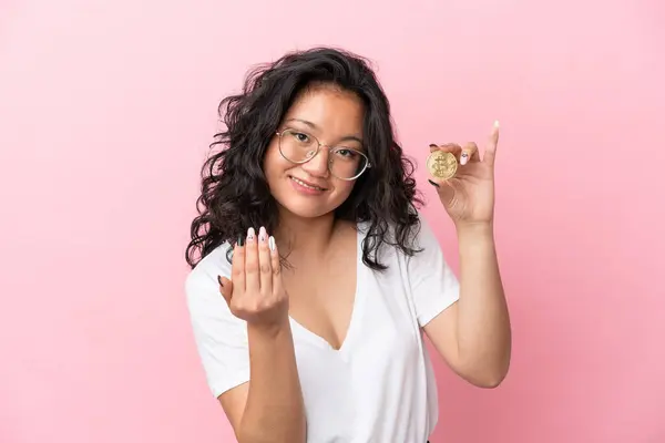 Young asian woman holding a Bitcoin isolated on pink background inviting to come with hand. Happy that you came