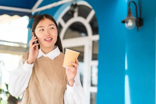 年轻的中国女人在户外用手机 带着愉快的表情喝咖啡 — 图库照片