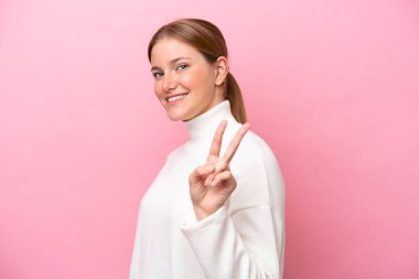 Pembe arka planda izole edilmiş genç beyaz kadın gülümsüyor ve zafer işareti gösteriyor.
