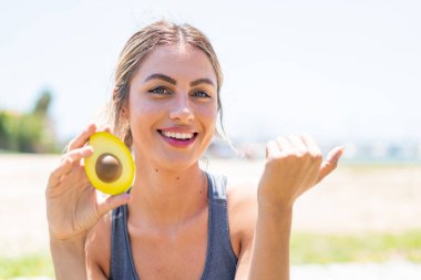 Avokado tutan genç sarışın bir kadın bir ürünü sunmak için yan tarafı işaret ediyor.