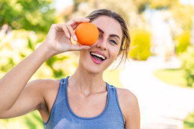 Açık havada genç sarışın bir kadın elinde mutlu bir ifadeyle bir portakal tutuyor.