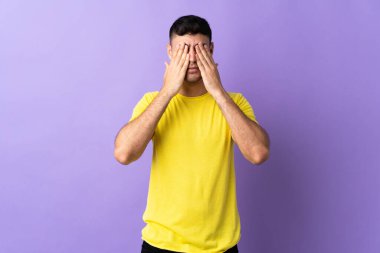 Mor arka planda izole edilmiş genç Kolombiyalı adam gözleri elleriyle kapatıyor.