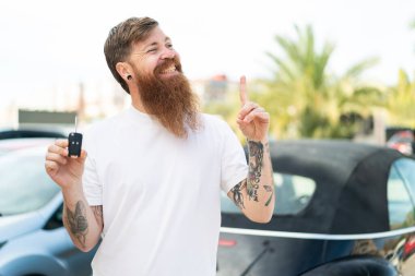 Dışarıda araba anahtarı tutan sakallı kızıl saçlı adam çözümü bulmak için parmağını kaldırıyor.
