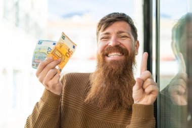 Kızıl saçlı sakallı adam dışarıda çok para alıyor ve harika bir fikri işaret ediyor.