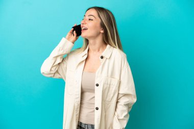 İzole edilmiş mavi arka planı olan genç bir kadın cep telefonuyla sohbet ediyor.