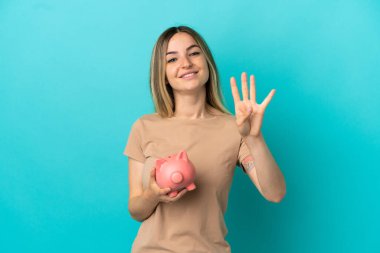 İzole edilmiş mavi arka planda kumbarayı tutan genç bir kadın mutlu ve parmaklarıyla dörde kadar sayıyor.