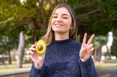 Genç, güzel Romen kadın avakado tutuyor, gülümsüyor ve zafer işareti gösteriyor.