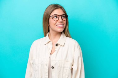 Genç beyaz kadın mavi arka planda izole edilmiş, mutlu bir ifadeyle gözlüklü.