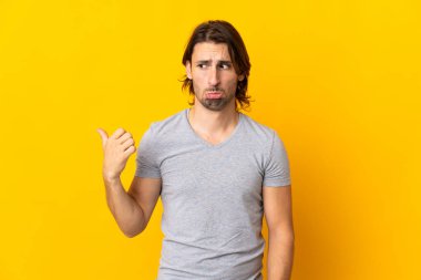 Sarı arka planda izole edilmiş genç ve yakışıklı bir adam mutsuz ve bir tarafı işaret ediyor.