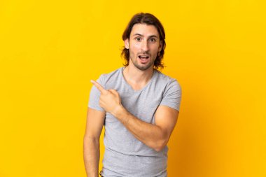 Sarı arka planda soyutlanmış yakışıklı genç adam şaşırmış ve işaret ediyor.