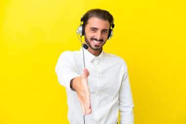 Tele pazarlamacı beyaz adam sarı arka planda izole edilmiş bir kulaklıkla çalışıyor ve iyi bir anlaşma için el sıkışıyor.