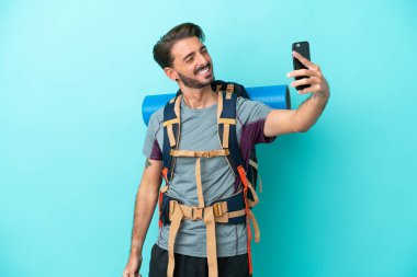 Mavi arka planda izole edilmiş büyük bir sırt çantası olan genç dağcı selfie çekiyor.