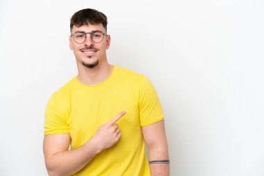 Beyaz arka planda izole edilmiş beyaz tenli yakışıklı genç bir adam bir ürün sunmak için kenarı işaret ediyor.