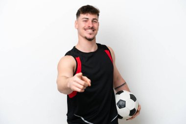 Beyaz arka planda, futbol topuyla izole edilmiş ve ön tarafı işaret eden beyaz tenli yakışıklı bir adam.