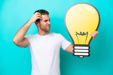 Mavi arka planda izole edilmiş genç beyaz bir adam elinde bir ampul simgesi tutuyor ve kuşkuları var.