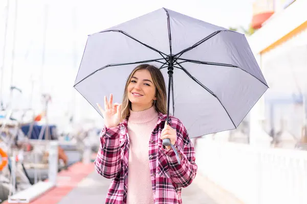 若い美しいルーマニアの女性は 指でOkのサインを示す屋外で傘を持っています — ストック写真