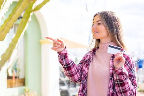 Young pretty Romanian woman holding a credit card at outdoors pointing to the side to present a product