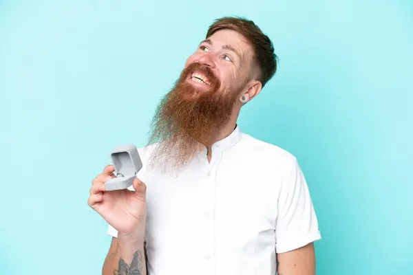 笑顔を見ながら青い背景に隔離された婚約指輪を保持する長いひげを持つ赤毛の男 — ストック写真