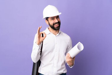 Miğferli genç bir mimar ve elinde iki eliyle onay işareti olan izole edilmiş mor arka plan planları var.
