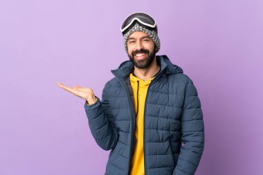 İzole edilmiş mor arka planda snowboard gözlüklü kayakçı bir reklam için fotokopi makinesi tutuyor.
