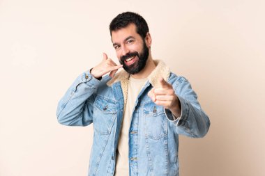 İzole edilmiş arka planda sakallı beyaz bir adam telefon hareketi yapıyor ve ön tarafı işaret ediyor.