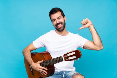 İzole edilmiş mavi arka planda gitar çalan genç bir adam gururlu ve kendini beğenmiş.