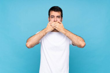 İzole edilmiş mavi arka planda sakallı genç bir adam elleriyle ağzını kapatıyor.