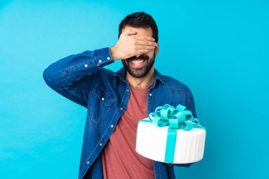 İzole edilmiş mavi arka planda büyük bir pastası olan genç yakışıklı adam gözlerini elleriyle kapatıyor.