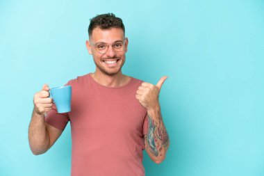 Kafkasyalı genç bir adam mavi arka planda izole edilmiş bir fincan kahve tutuyor ve bir ürünü sunmak için yan tarafı işaret ediyor.