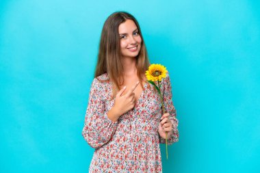 Mavi arka planda izole edilmiş ayçiçeği tutan beyaz bir kadın bir ürünü sunmak için kenarı işaret ediyor.