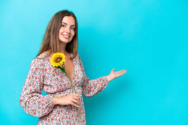 Mavi arka planda izole edilmiş ayçiçeği tutan genç beyaz kadın, davet edilmek için ellerini yana uzatıyor.