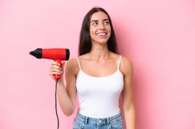 Kafkasyalı genç bir kadın pembe arka planda izole edilmiş saç kurutma makinesi tutuyor gülümserken yukarı bakıyor.