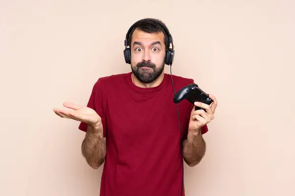 stock image Man playing with a video game controller over isolated wall having doubts while raising hands