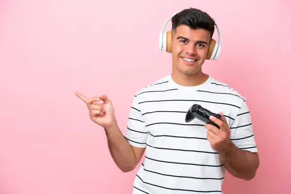 一个年轻英俊的男人正在玩一个用粉色背景隔离的电子游戏控制器 手指指向旁边 — 图库照片