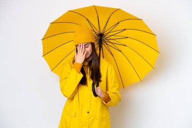 Yağmura dayanıklı ceketi ve şemsiyesi olan Brezilyalı genç bir kadın beyaz arka planda izole edilmiş, yorgun ve hasta bir ifadeyle.