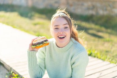 Genç, güzel bir kız, elinde sürpriz ve şok olmuş bir yüz ifadesiyle dışarıda hamburger tutuyor.