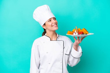 Kafkasyalı genç aşçı kadın elinde waffle tutarken mavi arka planda izole edilmiş gülümserken yukarı bakıyor.