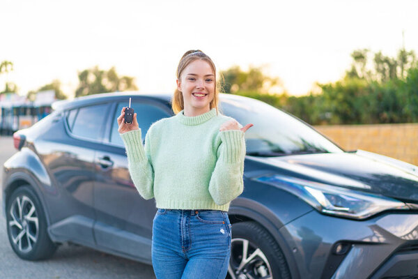 Young pretty girl holding car keys at outdoors pointing to the side to present a product