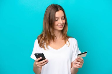 Genç beyaz kadın mavi arka planda izole edilmiş cep telefonuyla kredi kartıyla alışveriş yapıyor.