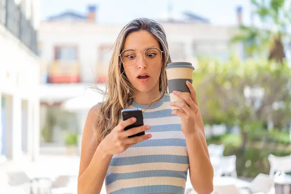 Young Pretty Woman Outdoors Using Mobile Phone Holding Coffee Surprised — Stock Photo, Image