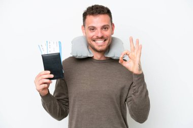 Beyaz arka planda pasaport taşıyan genç beyaz bir adam parmaklarıyla tamam işareti gösteriyor.