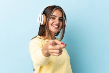 Genç Uruguaylı kadın mavi arka planda izole edilmiş müzik dinliyor ve ön tarafı işaret ediyor.